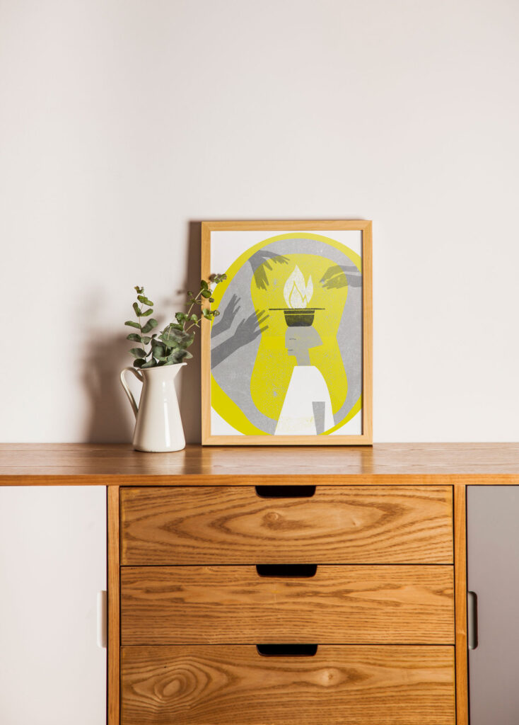 Un aparador de madera con una jarra blanca que contiene vegetación y una obra de arte abstracta enmarcada que muestra manos alrededor de una antorcha encendida con un fondo amarillo, contra una pared blanca sencilla.